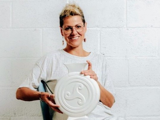 sobre fondo blanco, la artesana Eva Conde vestida de blanco sujeta una de sus piezas de cerámica blanca con relieve de un motivo celta