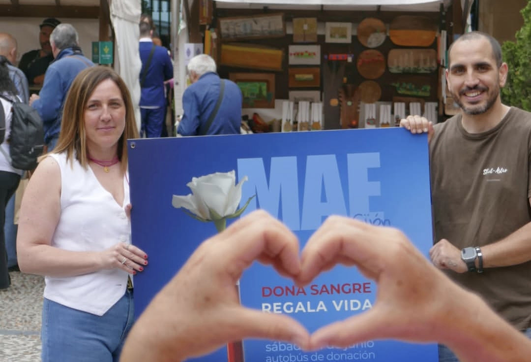 dos personas sujetan el cartel de la campaña de donación de sangre en el MAE, al fondo el mercado, y frente al cartel unas manos hacen al juntarse el símbolo del corazon 
