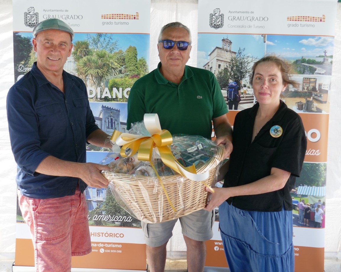 tres personas posan frente al lote de productos que forma parte de los premios de la actividad sobre el camino primitivo en el mae
