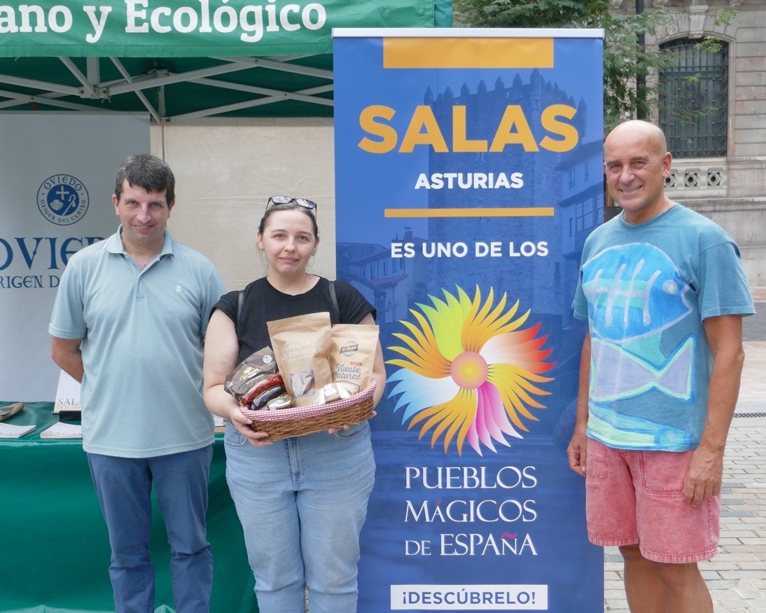 tres personas posan frente al lote de productos que forma parte de los premios de la actividad sobre el camino primitivo en el mae