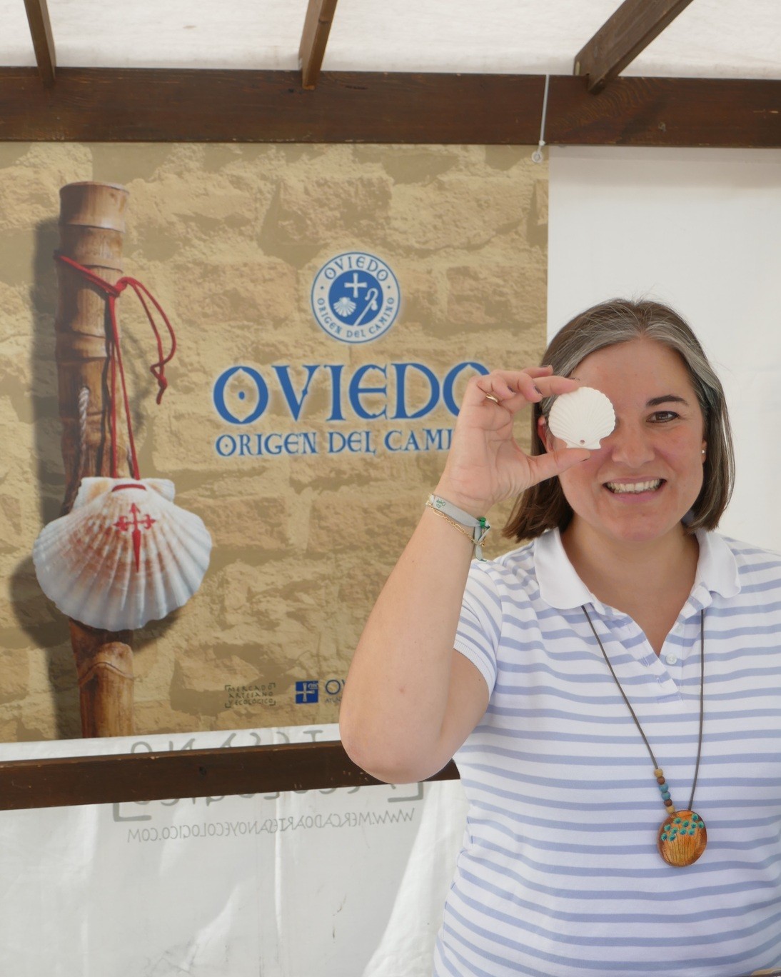 una mujer frente al cartel de oviedo origen del camnio posa con un colgante de cerámica con forma de concha colocado sobre su ojo