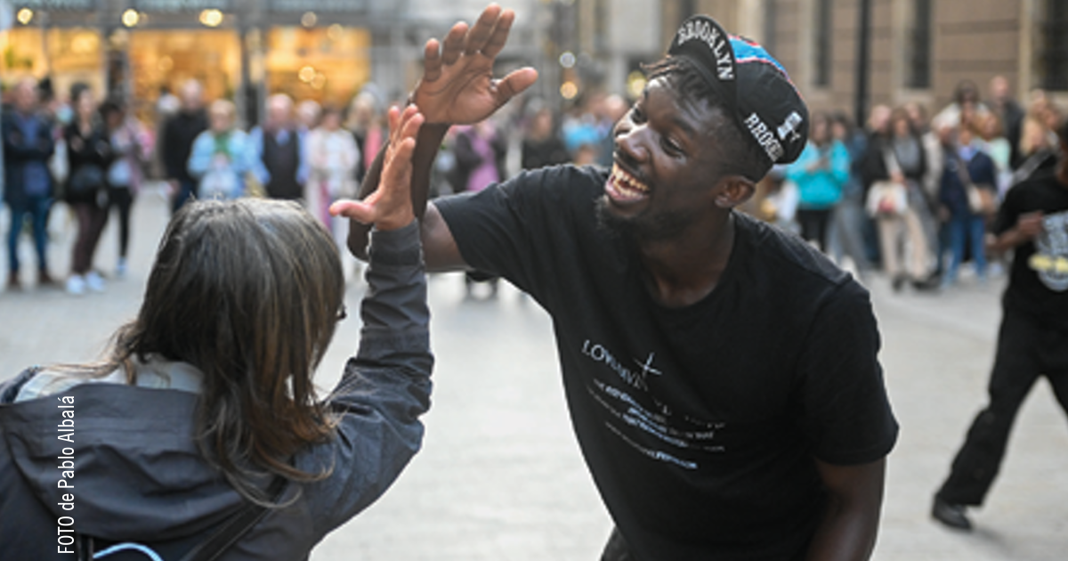 Dos personas chocando las manos alegremente en la calle; un hombre sonriente con camiseta negra y gorra interactúa de cerca con otra persona de espaldas, frente a una multitud difuminada en un entorno urbano