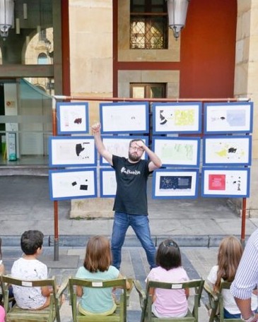 Animador narrando un cuento al aire libre ante varios niños, con una exposición de ilustraciones enmarcadas al fondo.
