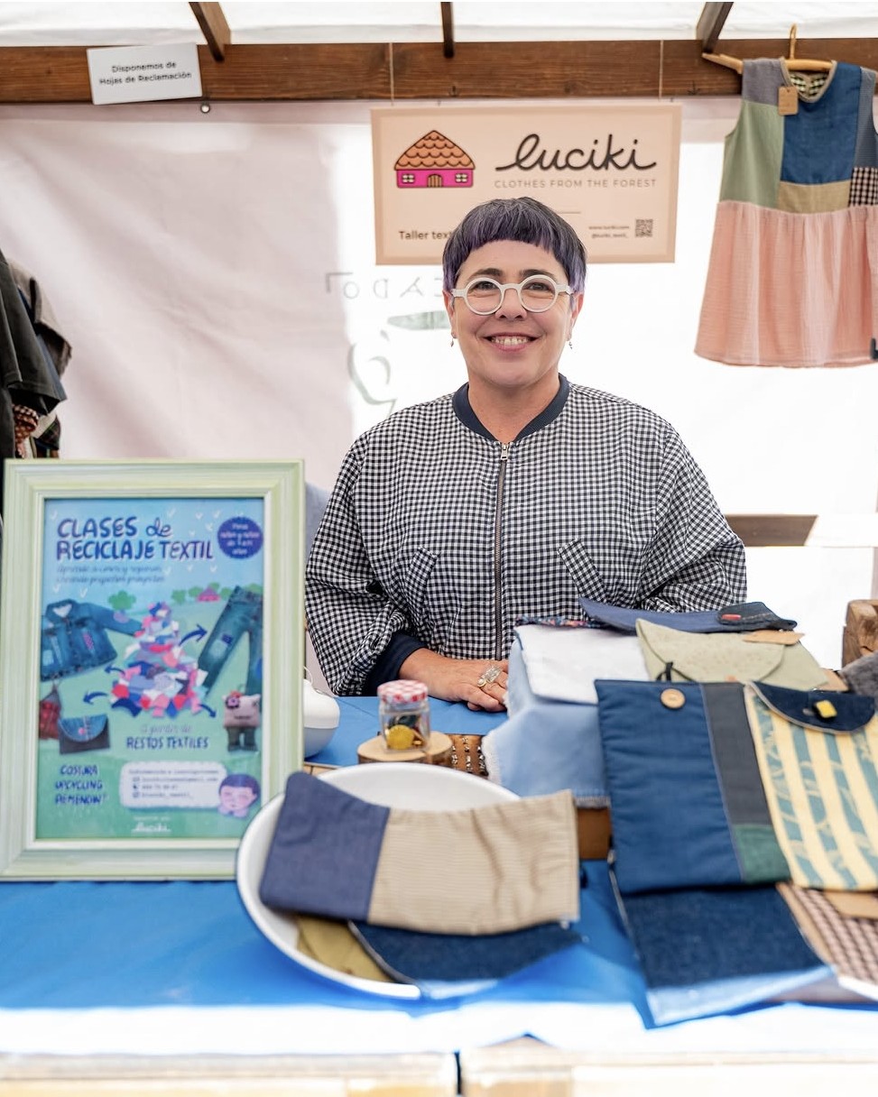 Una artesana sonríe detrás del mostrador de su puesto llamado luciki, rodeada de prendas de ropa y accesorios textiles de diseño sostenible.