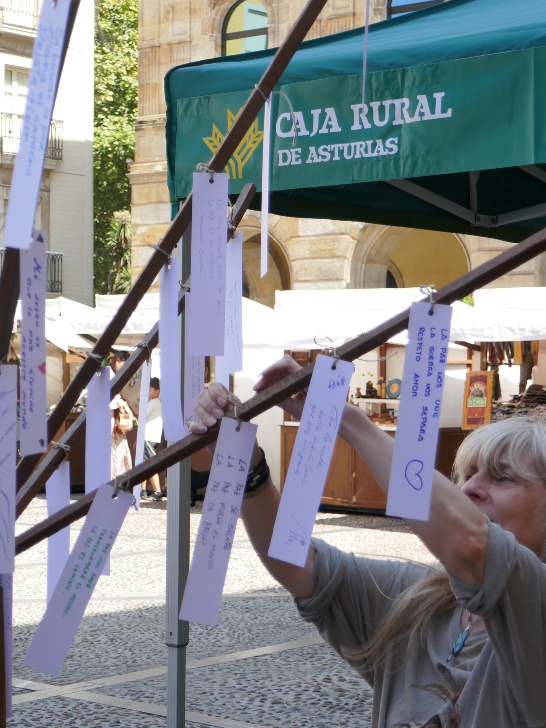Una mujer, de perfil, coloca con cuidado un pequeño papel manuscrito, que contiene un haiku, con una pinza decorativa en una de las ramas de un árbol frondoso situado dentro del recinto del Mercado Artesano y Ecológico de Gijón (MAE). El árbol está lleno de otros haikus similares ya colgados por los visitantes, creando un ambiente poético y participativo.