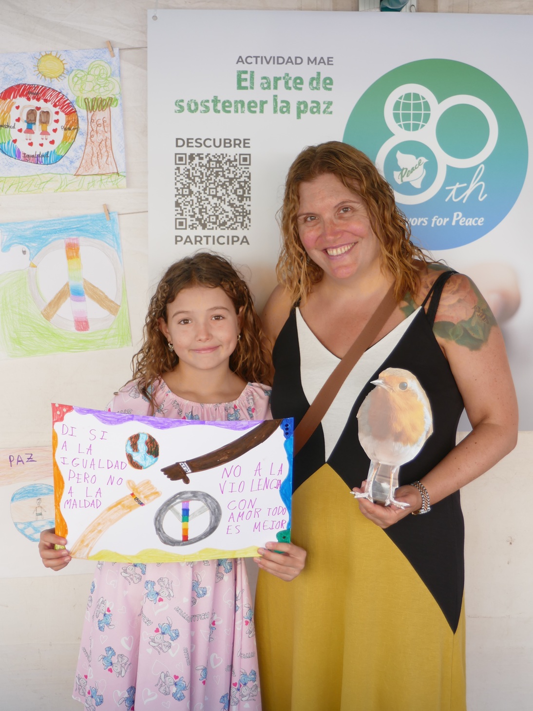 Primer plano de una madre y su hija sonrientes en el Mercado Artesano y Ecológico de Gijón (MAE). La niña muestra orgullosa un dibujo hecho por ella misma con colores vivos, el cual lleva una pequeña etiqueta o sello que lo identifica como su participación en el Concurso Infantil del Día de la Paz del MAE. Ambas miran a cámara con alegría.