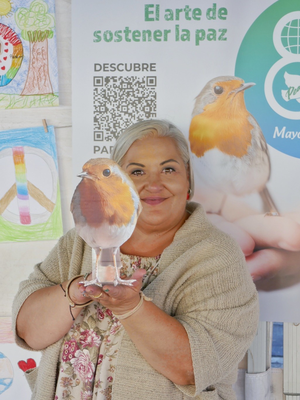 Plano medio de una artesana del Mercado Artesano y Ecológico de Gijón (MAE), sosteniendo tiernamente un pequeño petirrojo posado sobre la palma de sus manos abiertas. Detrás de ella, en la pared del puesto, se puede ver claramente el cartel oficial del MAE para el Día de la Paz, que reproduce la misma imagen icónica del petirrojo sobre las manos.