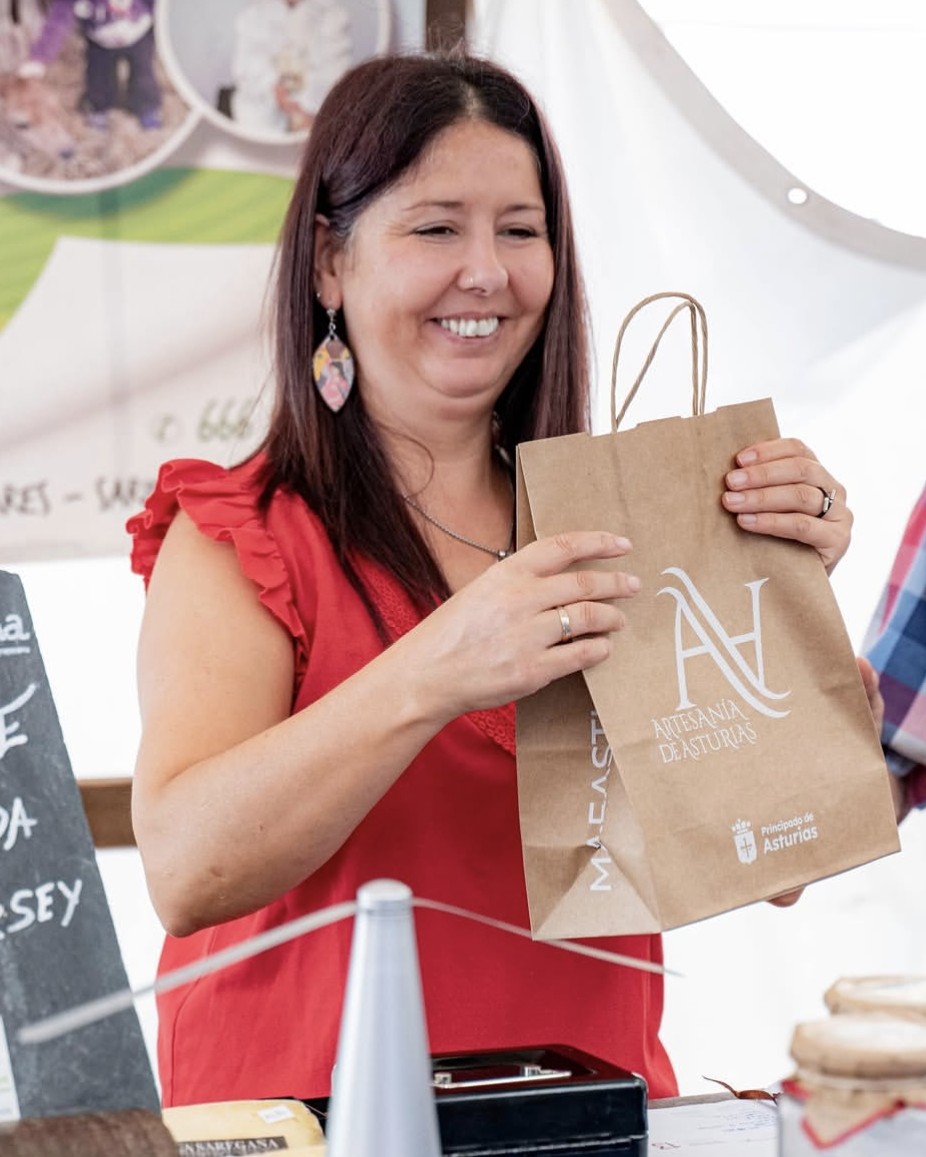Una mujer sonriente con blusa roja sostiene una bolsa de papel marrón con el logotipo de "Artesanía de Asturias" en un puesto del mercado artesano y ecologico