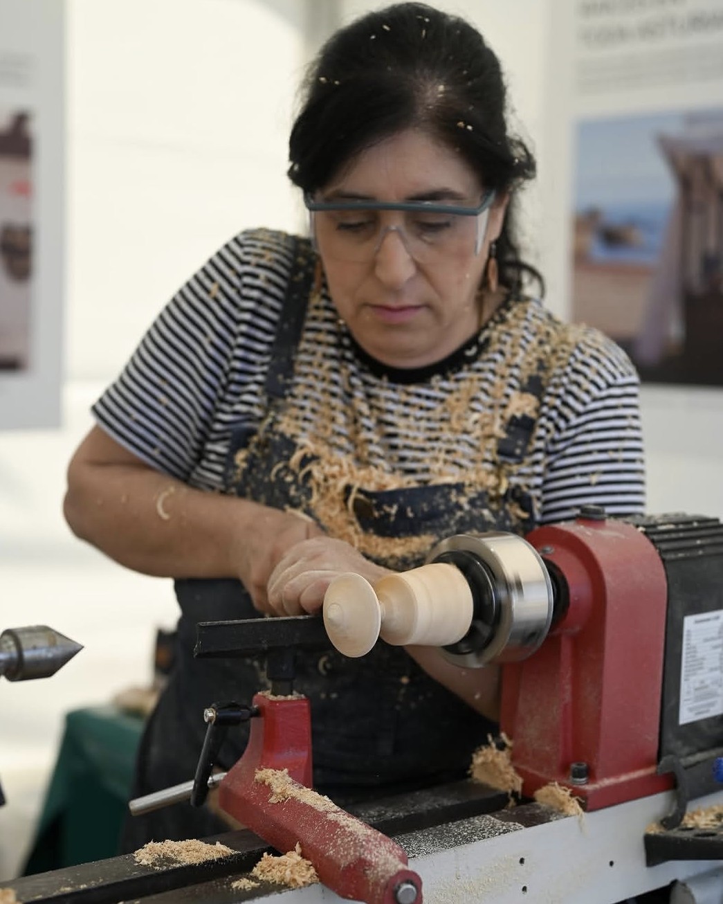 Una mujer artesana con gafas de protección y delantal trabaja la madera en un torno mecánico, modelando una pieza mientras se aprecian virutas a su alrededor.